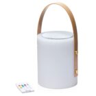 A cylindrical portable speaker with a white body and a tan leather handle. It is standing upright with a small remote control placed next to it on a white surface. The remote has colorful buttons.