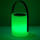 A cylindrical, glowing green lantern with a black handle emits a soft light in a dark setting. The lantern has a modern design, creating a luminous and ambient atmosphere.