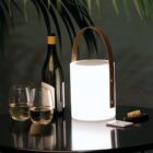 A softly glowing, portable lantern with a leather handle sits on a dark table next to an open wine bottle and two filled glasses. A cork and a small remote control are nearby. Large leaves hang overhead, casting shadows.