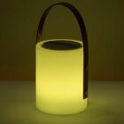 A glowing cylindrical lamp with a smooth, opaque surface emits a warm light. It has a sleek, dark handle attached to the sides, creating a modern, portable design. The background is solid and dark, enhancing the lamps illumination.