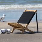 A wooden beach chair with a black fabric seat sits on sandy shore near the ocean. Two rolled towels and a pair of sandals are beside it. A seagull walks along the waters edge in the background, with gentle waves approaching.