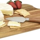 A wooden serving board with a knife, sliced brie cheese on crackers, and whole brie cheese. Two strawberries are also placed on the board.