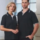 Two people wearing matching black polo shirts with white trim stand in a bright room. The woman has shoulder-length blonde hair, and the man has short brown hair and a beard. They stand close together, looking at the camera.