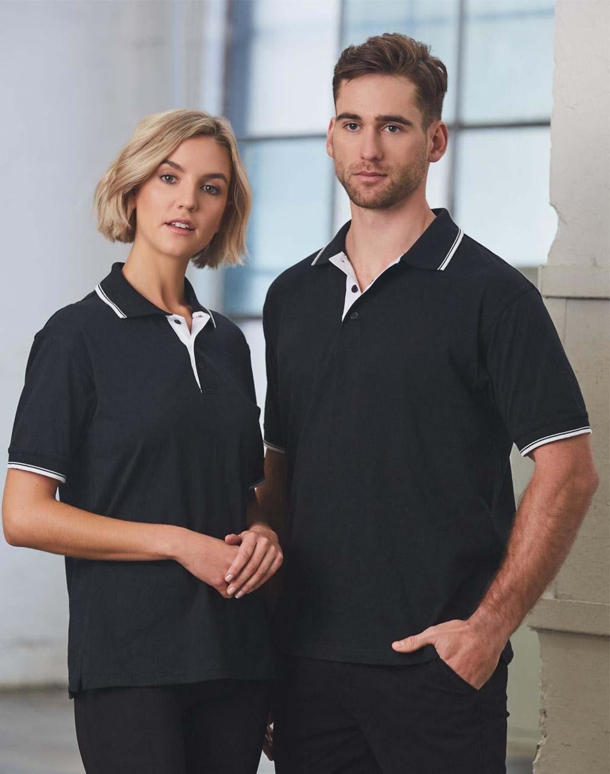 Two people wearing matching black polo shirts with white trim stand in a bright room. The woman has shoulder-length blonde hair, and the man has short brown hair and a beard. They stand close together, looking at the camera.