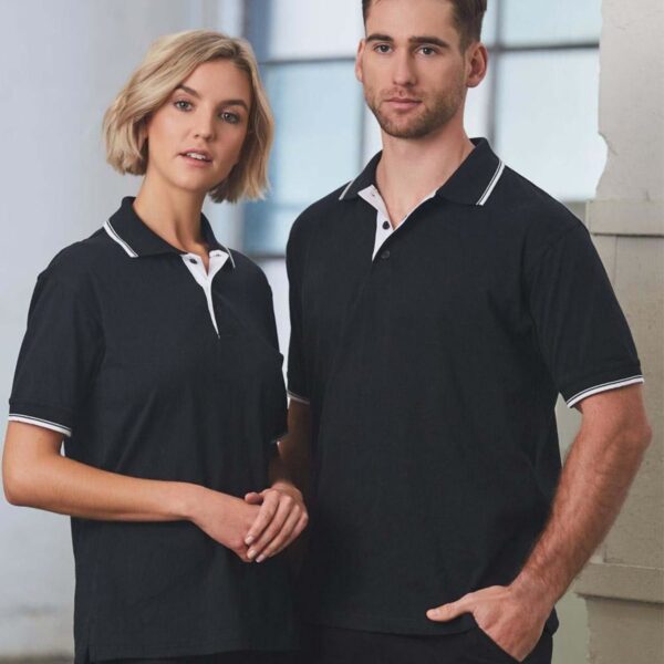 A woman and a man stand side by side, both wearing matching black polo shirts with white collars and trim. They are indoors, in front of a large window, and appear relaxed and composed.