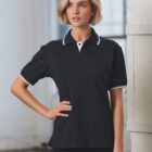 A person with short blonde hair wears a black polo shirt with white trim, standing indoors with a hand on their hip. They look to the side, next to a large window that lets in natural light.