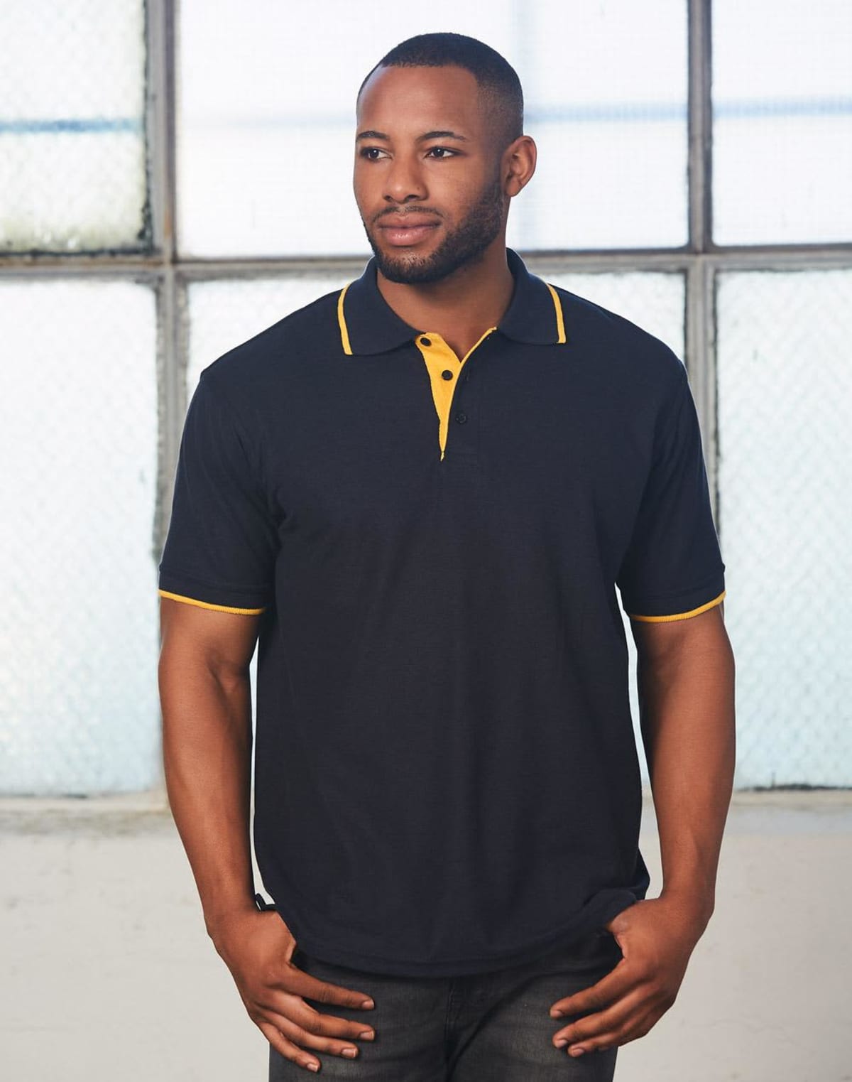 A person wearing a black polo shirt with yellow trim stands in front of a large, industrial-style window. The person looks to the side with a neutral expression and has short hair.
