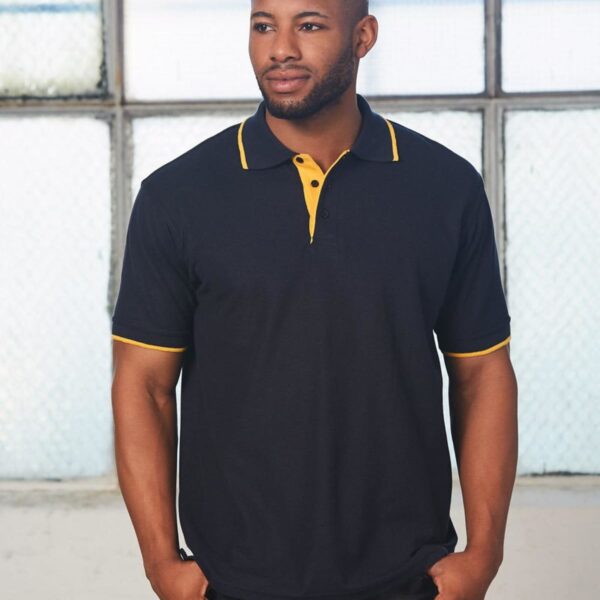 A man wearing a black polo shirt with yellow trim stands indoors in front of a large window. The room has a soft, diffused light, and the man is looking slightly to the side.