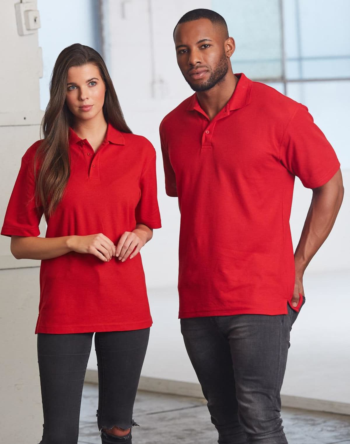 A woman and a man are standing side by side, wearing matching red polo shirts. The woman has long brown hair and is wearing jeans. The man has short hair and is also wearing jeans. They are standing in a bright, neutral indoor space.