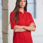 Woman with long brown hair wearing a red polo shirt and black pants stands with arms crossed in a brightly lit room with white walls and large windows.