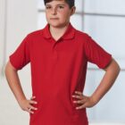 A boy stands confidently with hands on hips, wearing a red polo shirt and blue jeans. The background is a softly lit white interior with large window panes.