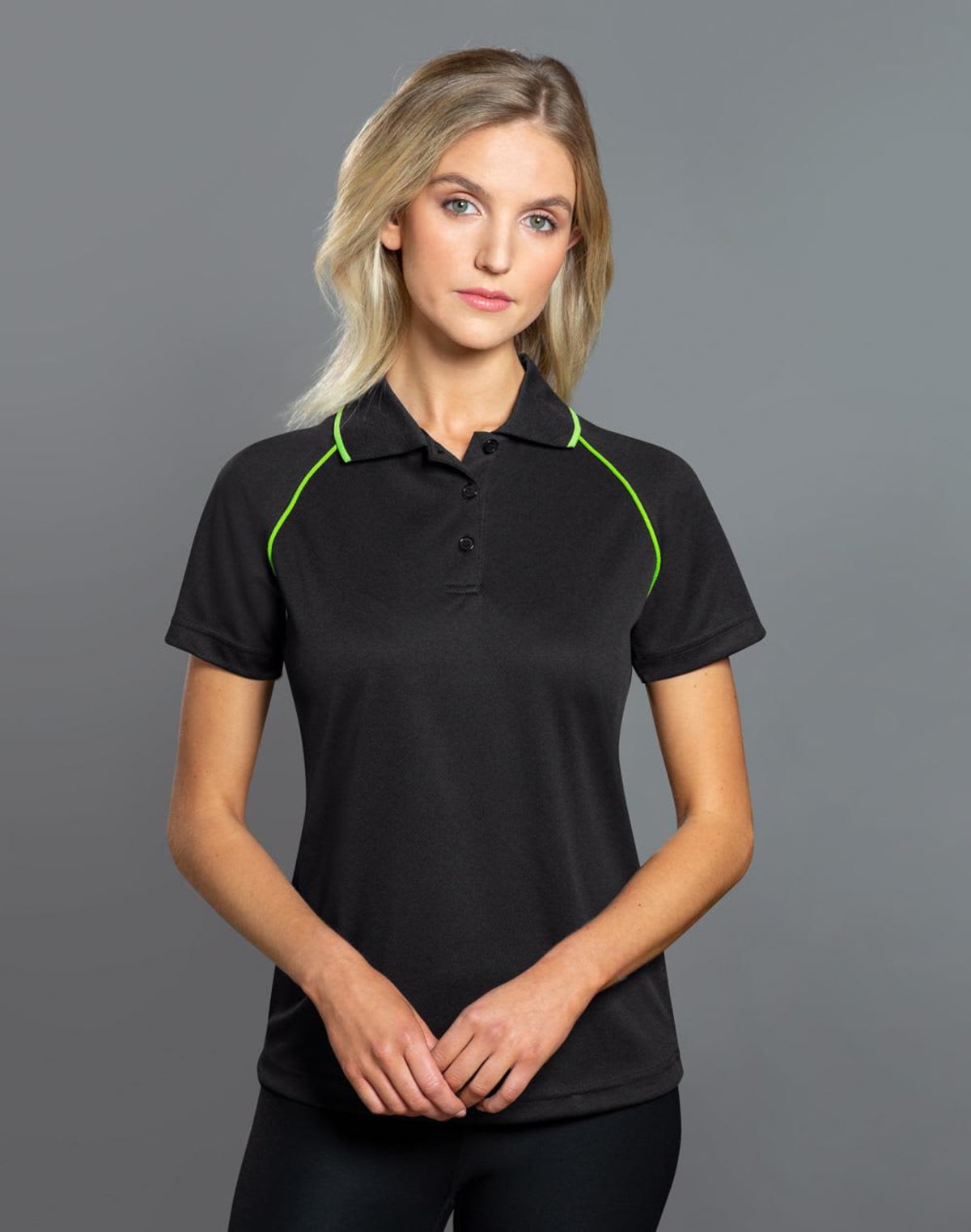 A person with long blonde hair wearing a black polo shirt with green piping stands against a gray background, looking forward with hands clasped.
