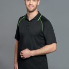 A person wearing a black polo shirt with green accents stands against a gray background. They have short hair and are smiling while looking at the camera, with one hand loosely crossed over their other wrist.