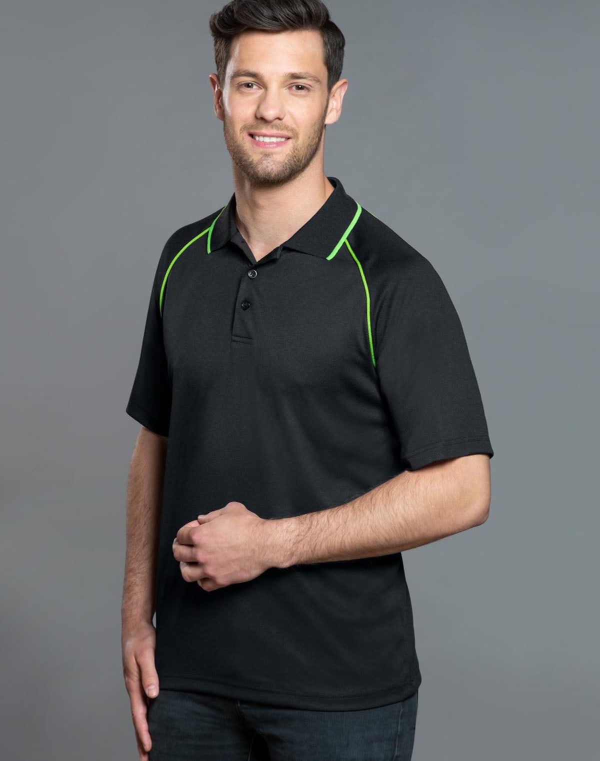 A person wearing a black polo shirt with green accents stands against a gray background. They have short hair and are smiling while looking at the camera, with one hand loosely crossed over their other wrist.