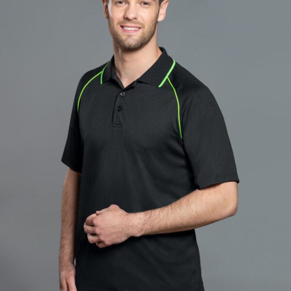 A man wearing a black polo shirt with green piping on the shoulders stands against a gray background. He has short hair and a beard, and he is smiling with one hand resting near his waist.