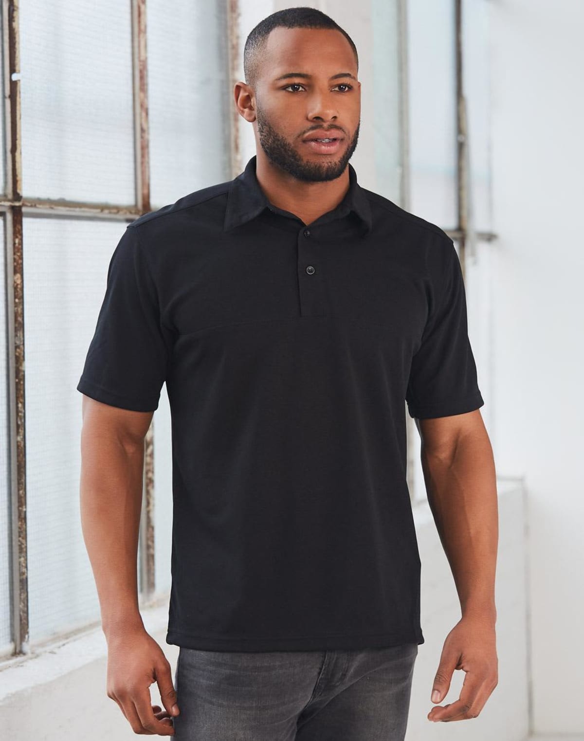 A man wearing a black polo shirt and gray jeans stands indoors near large windows, looking slightly to the side. The room has white walls and natural light coming through the windows.