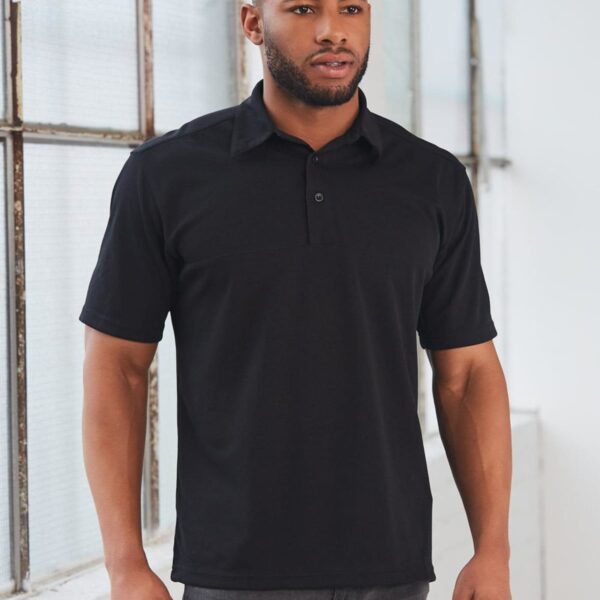 Man wearing a black polo shirt and jeans stands indoors near large windows, looking in the distance. The setting is well-lit, emphasizing the casual and modern style of his outfit.