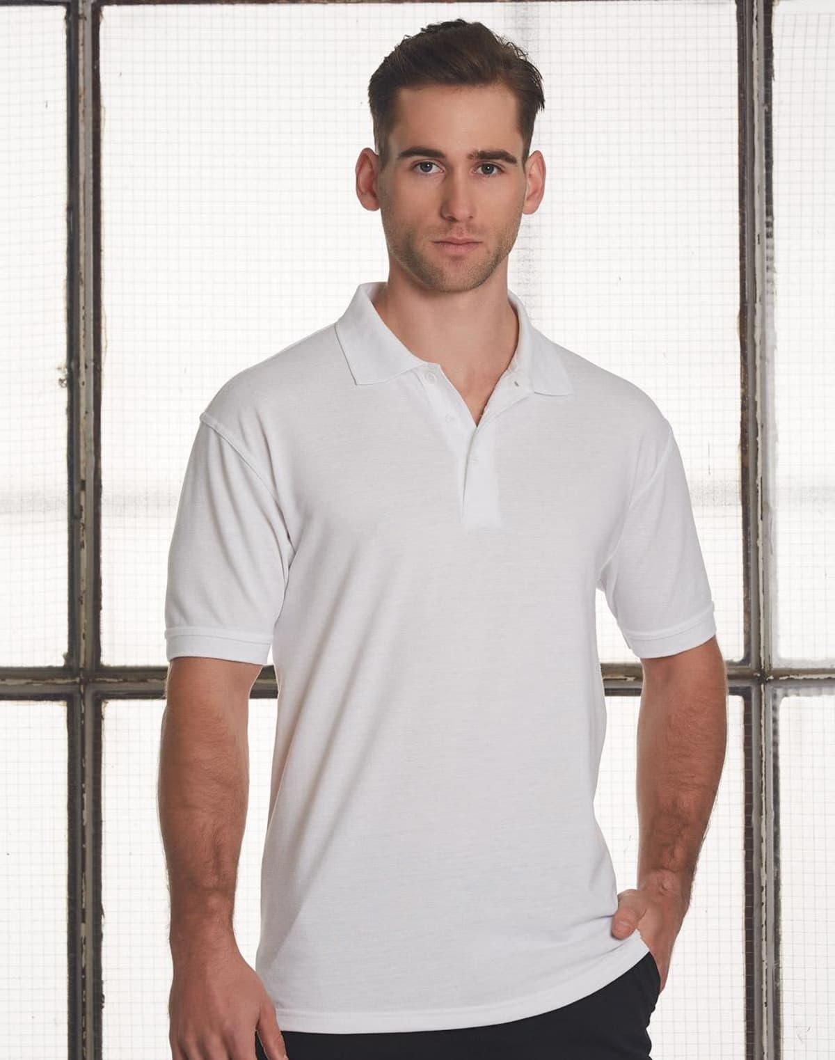 A man wearing a white polo shirt stands against a large, grid-patterned window. He has short hair and a neutral expression, with his left hand resting on his hip. The background is softly lit.