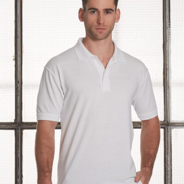 A man wearing a white polo shirt stands in front of a large window with a grid pattern. He looks directly at the camera with a neutral expression, and his hands are relaxed by his sides.