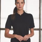 A person with long dark hair is standing in front of a window, wearing a black polo shirt. The background is bright, and the window has a grid pattern. The persons hands are gently clasped in front.