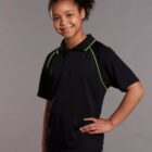 A young person with curly hair smiles while wearing a black polo shirt with green piping. They have one hand on their hip and stand against a plain gray background.