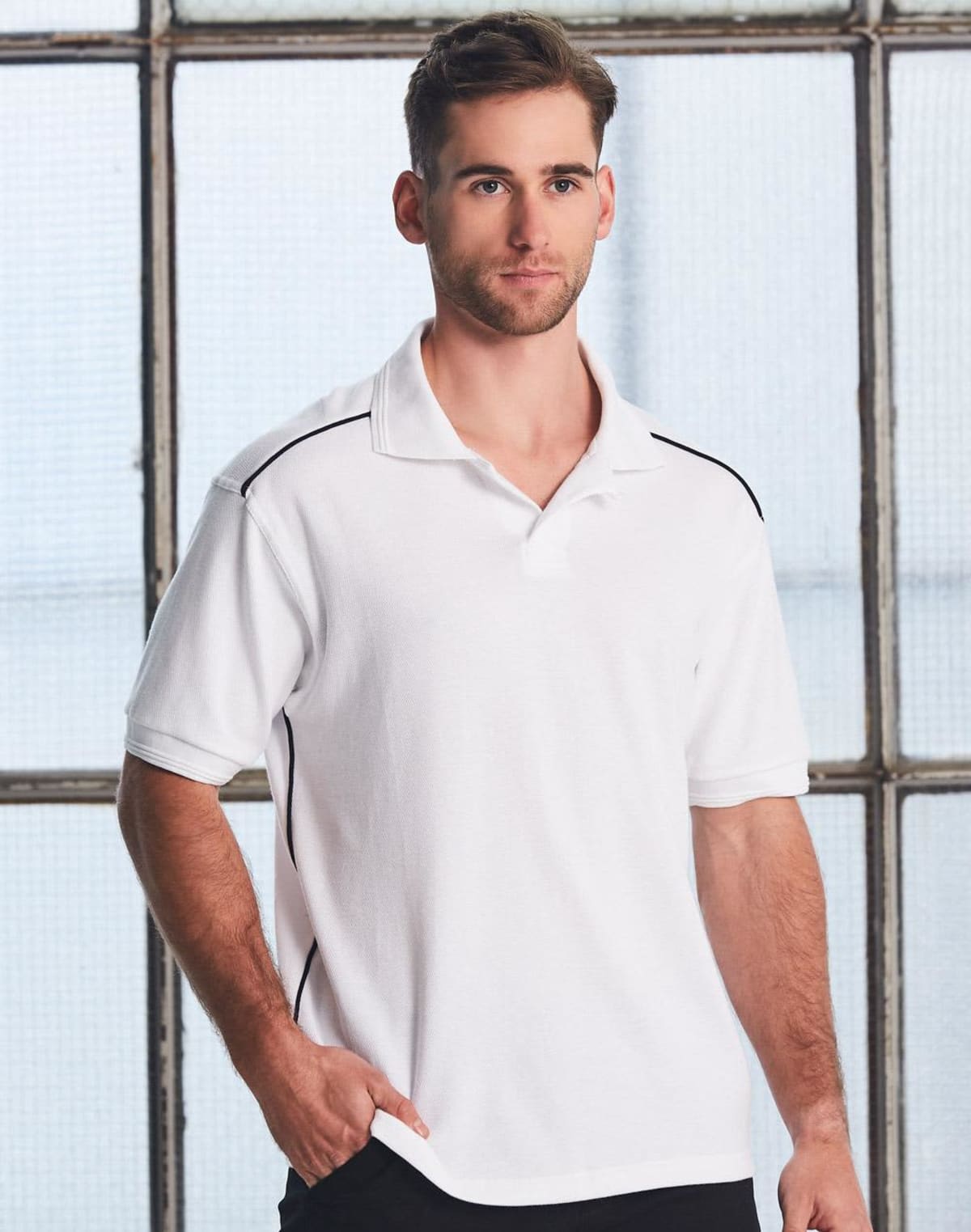 A person wearing a white polo shirt with black trim stands in front of a large window with grid patterns. They have short hair and a beard, appearing relaxed with one hand in their pocket.