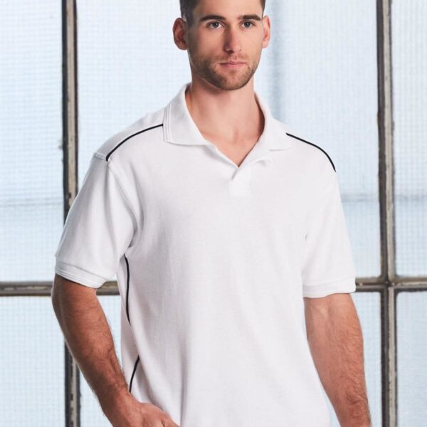 A man with short hair is wearing a white polo shirt with black trim along the shoulders and sides. He stands in front of a large industrial-style window with a grid design.