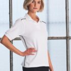 A person with short blonde hair is posing in a white polo shirt with black piping, standing against a large frosted window. They have one hand on their hip and a neutral expression.