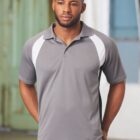 A person stands indoors wearing a gray polo shirt with white side panels. The background features blurred architectural elements and a window.