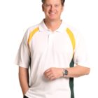 A smiling man stands with one hand in his pocket, wearing a white polo shirt with yellow and green accents and dark pants. He has short brown hair and is wearing a watch on his left wrist. The background is plain white.
