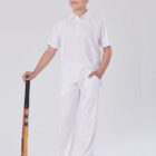 A young boy stands confidently holding a cricket bat. Hes dressed in all-white sports attire, including a polo shirt and trousers, on a plain white background. His stance is relaxed, with a slight smile on his face.