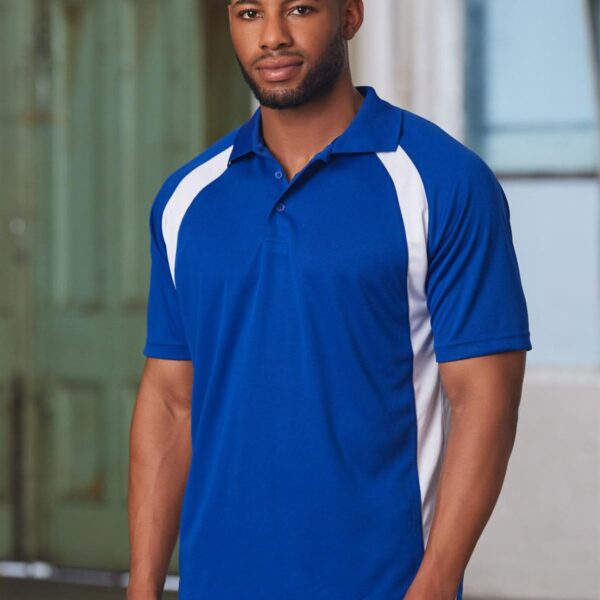 A man wearing a blue and white polo shirt stands indoors in front of a blurred background featuring a green door and a window. He has short hair and a beard, and hes looking directly at the camera.