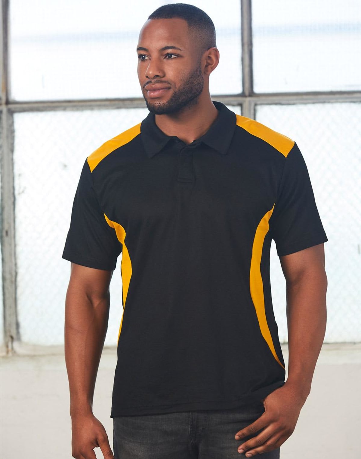 A person wearing a black polo shirt with yellow accents stands indoors in front of a window, looking to the side.