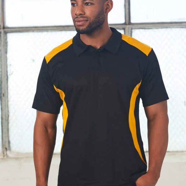 A person wearing a black polo shirt with yellow accents stands indoors by large windows, looking to the side.
