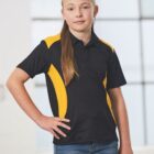 A young girl with long blond hair stands confidently wearing a black and yellow sports polo shirt and blue jeans. She has a neutral expression and stands in a well-lit indoor setting with a large window in the background.