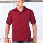 A person in a red polo shirt leans against a white surface, with arms resting on it. They have short, dark hair and are wearing gray pants. The background features a wire mesh panel and a white wall.