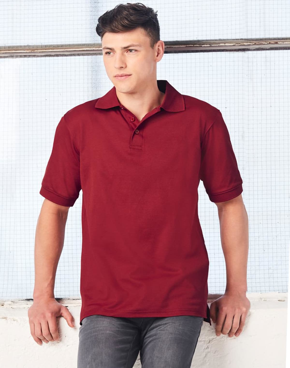 A person in a red polo shirt leans against a white surface, with arms resting on it. They have short, dark hair and are wearing gray pants. The background features a wire mesh panel and a white wall.