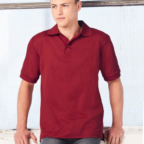 Person wearing a red polo shirt and gray pants, leaning against a white wall with a grid-patterned window in the background.