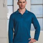 A person wearing a long-sleeve blue polo shirt stands indoors with one hand on their hip. They are looking directly at the camera. The background features large windows and part of a white wall.