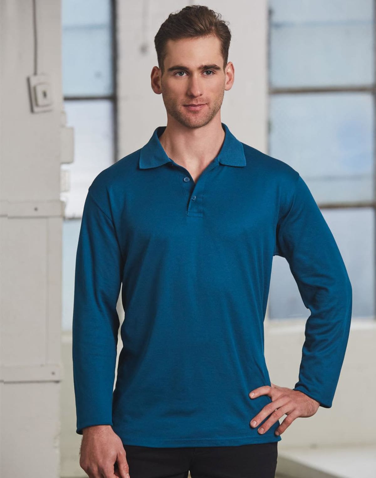 A person wearing a long-sleeve blue polo shirt stands indoors with one hand on their hip. They are looking directly at the camera. The background features large windows and part of a white wall.