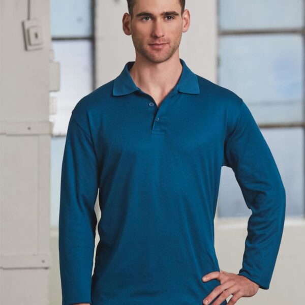 A person with short brown hair is standing indoors, wearing a long-sleeve teal polo shirt and black pants. They have one hand on their hip and are looking at the camera. The background features large windows and white walls.