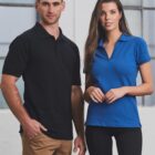 A man in a black polo shirt and brown pants stands next to a woman in a blue polo shirt and black pants. They are both looking at the camera, with a neutral expression. The background features large windows.