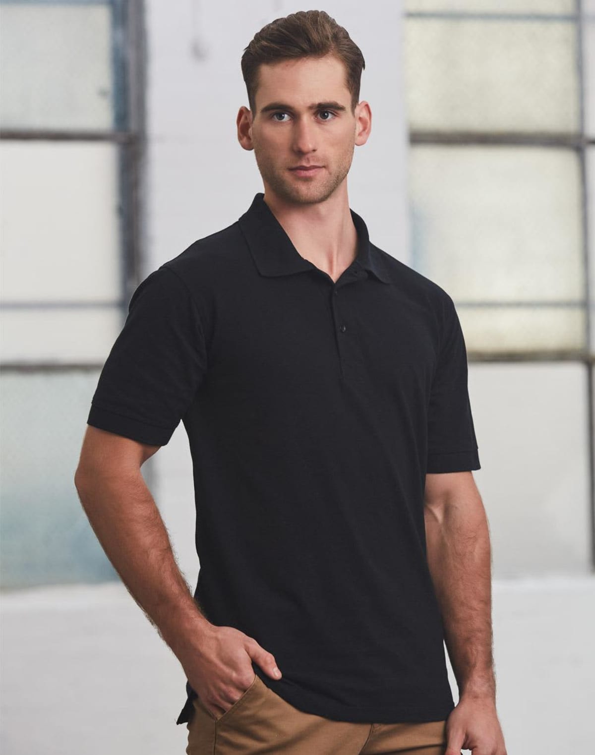 Man in a black polo shirt and brown pants standing in a well-lit room with large windows in the background. He has short brown hair and is looking confidently at the camera.