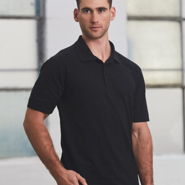 Man in a black polo shirt and brown pants stands indoors with industrial-style windows in the background. He has short brown hair and a neutral expression, with one hand resting on his hip.