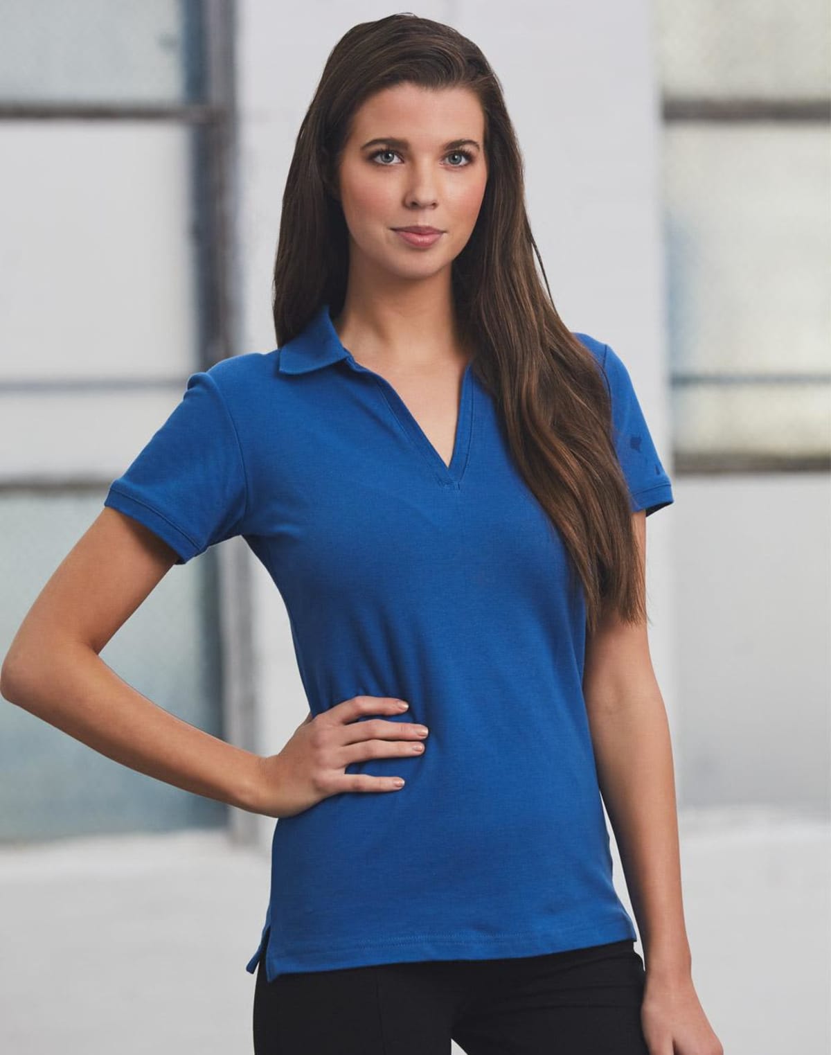 A woman with long brown hair wearing a blue polo shirt and black pants stands with one hand on her hip. She is in front of a blurred background with large windows.
