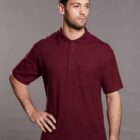 A man wearing a burgundy short-sleeve polo shirt with a pocket stands against a gray background. He has short dark hair and a beard, with one hand resting on his hip.
