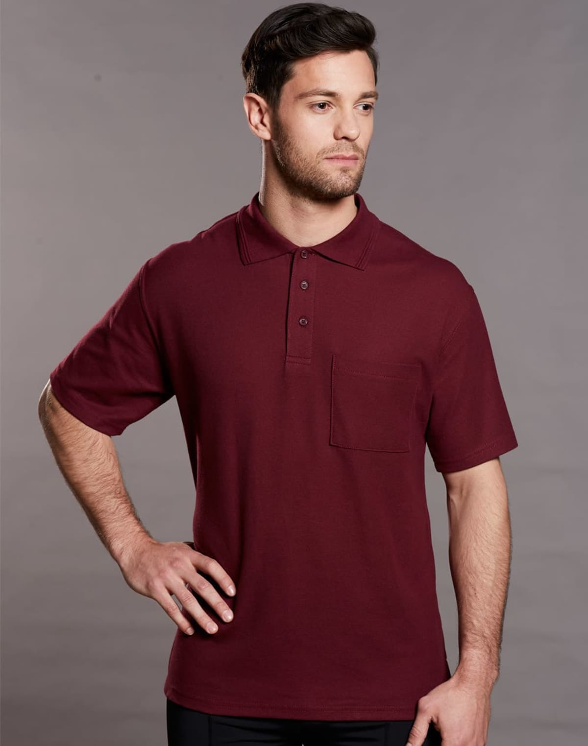 A man wearing a burgundy short-sleeve polo shirt with a pocket stands against a gray background. He has short dark hair and a beard, with one hand resting on his hip.