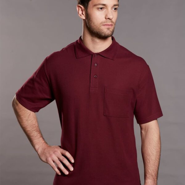 A person wearing a burgundy polo shirt poses against a plain gray background. The polo has a front pocket and buttoned collar. The person has one hand on their hip, looking slightly to the side.