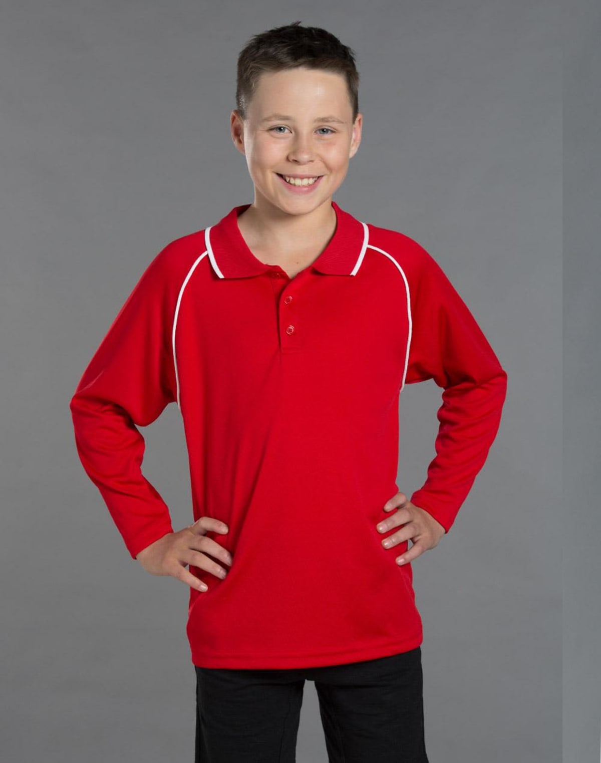 Smiling boy in a red long-sleeve polo shirt with hands on hips against gray background.