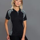 A woman with long blonde hair wearing a black and gray athletic polo shirt and black pants poses against a gray background. She is leaning on a white object, looking directly at the camera.